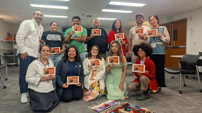 Group of community leaders holding orange flyers in a meeting room, standing behind a display of cultural textiles on floor.